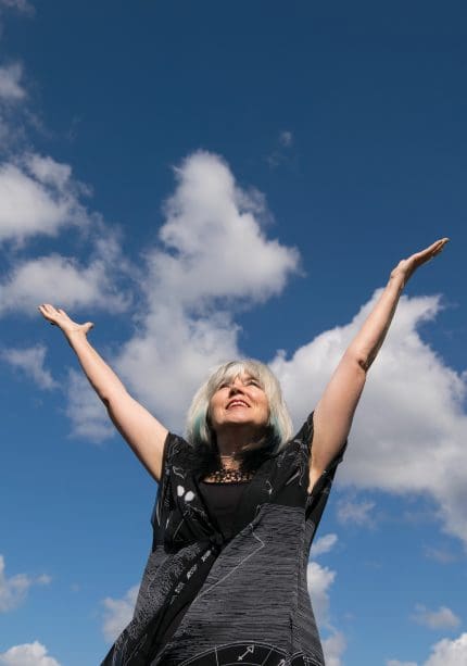 Faye Blake standing outdoors with both arms raised in a V shape against a bright blue sky with scattered clouds.