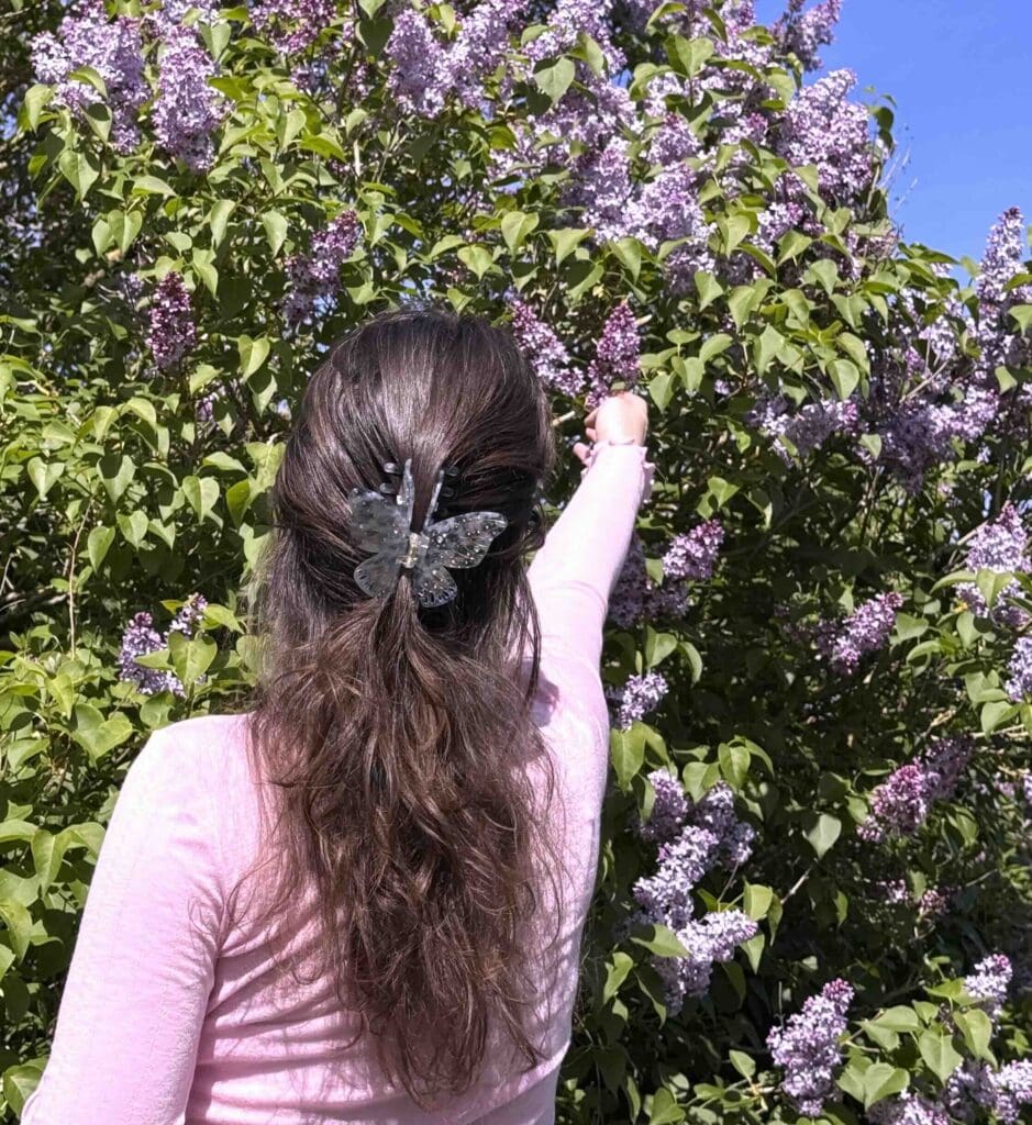 Nynne Noel reaching toward blooming lilac flowers in her garden, illustrating lived experience, seasonality, and embodied presence as part of place-based astrological practice in Denmark.